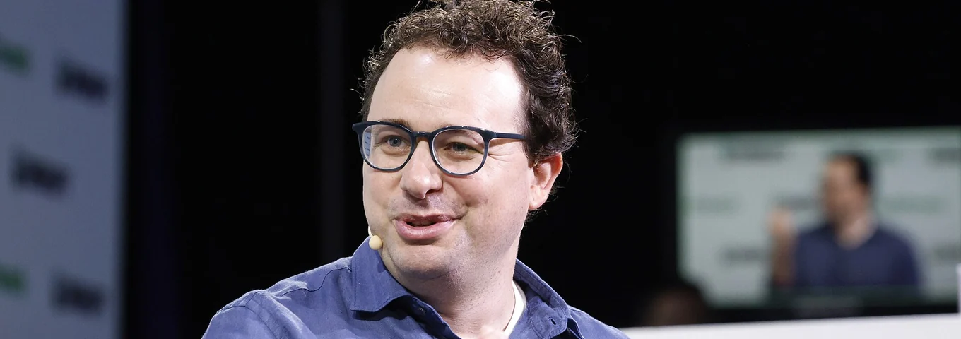 Dario Amodei in glasses gesturing with one hand as he speaks on stage at TechCrunch Disrupt, against a dark background.