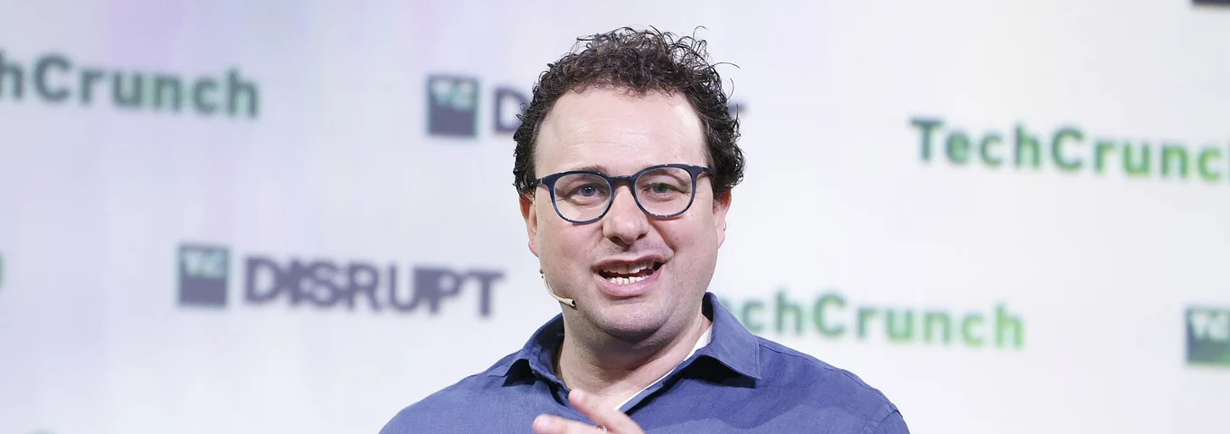 Dario Amodei, with curly dark hair, glasses and a blue shirt, speaking into a microphone on stage at TechCrunch Disrupt 2023.